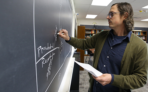 Male instructor standing at a chalk board writing a math problem. 