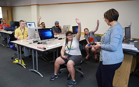 Students sitting in a compute lab raising their hands to be chosen by the instructor