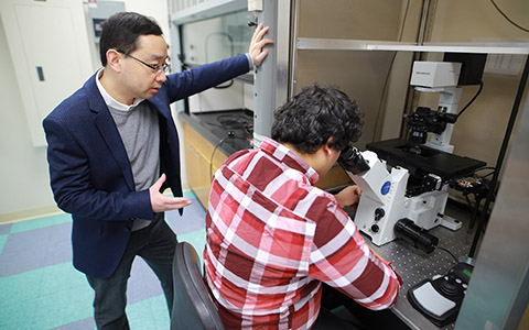A man stands and watches as another man looks through a microscope in a lab