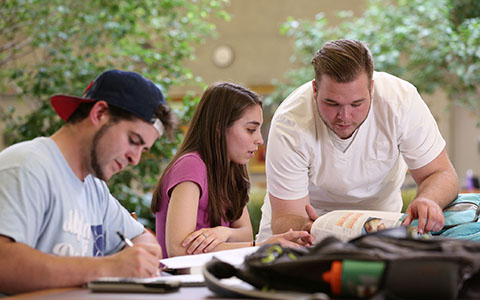 Three students at a table , two of them are looking at the same textbook togther the third student is writing things down on paper