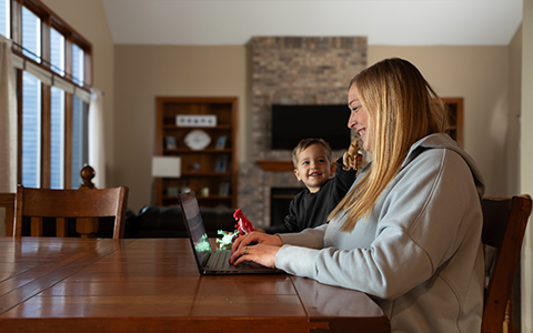 Woman working on a laptop at a table while her son is next to her