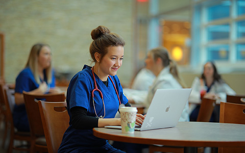 Man wearing blue nursing scrubs sits at a table with an open laptop