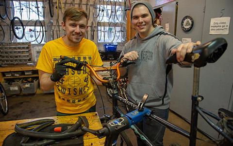 Two students smile and stand together in a repair workshop