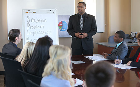 Man stands in front of group at a conference table and presents information on white board