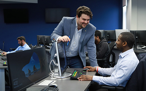 Student in a computer lab in front of a computer screen talking to another student