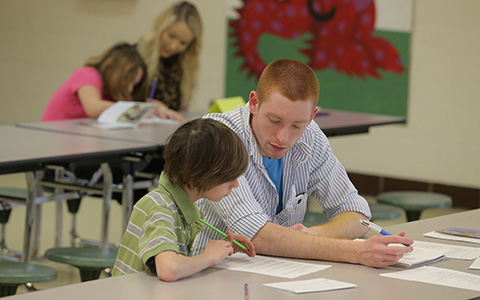 A man sits besides a child at a table in a classroom as the both look over one of the worksheets