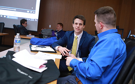 Two sales students dressed in suits sit at a table to talk with notes open