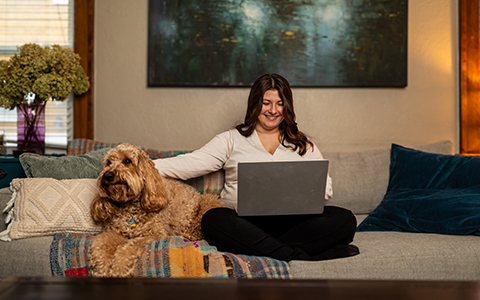 A woman sitting on a couch working on a laptop with her dog next to her