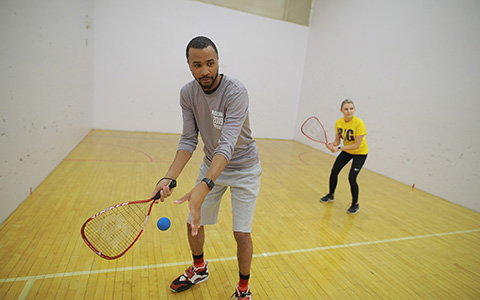 A man and woman in an indoor racquetball court prepare to serve the ball