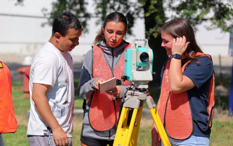 trio of students wearing orange vests surround a surveying camera to collect data