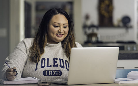 Woman wearing a UToledo sweatshirt looks down at her open laptop next to her pen and notebook